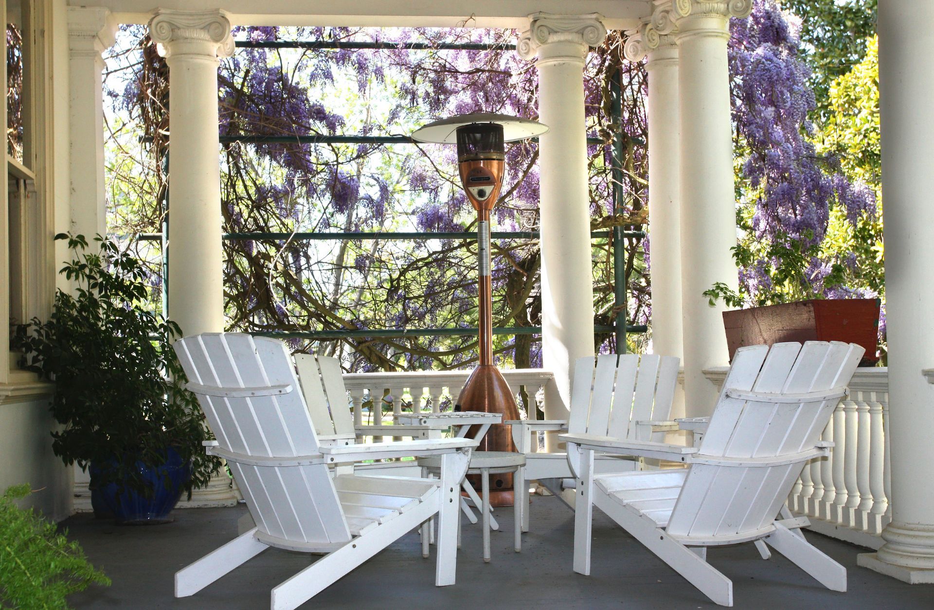 White Adirondack chairs around a table on a porch, with a heater and flowering vines.