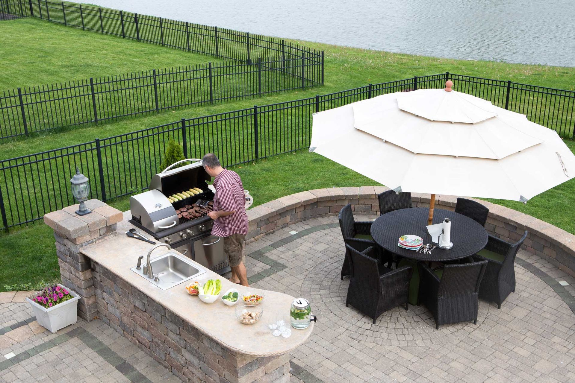 Man grilling food on outdoor kitchen, dining table with umbrella on patio overlooking lake.