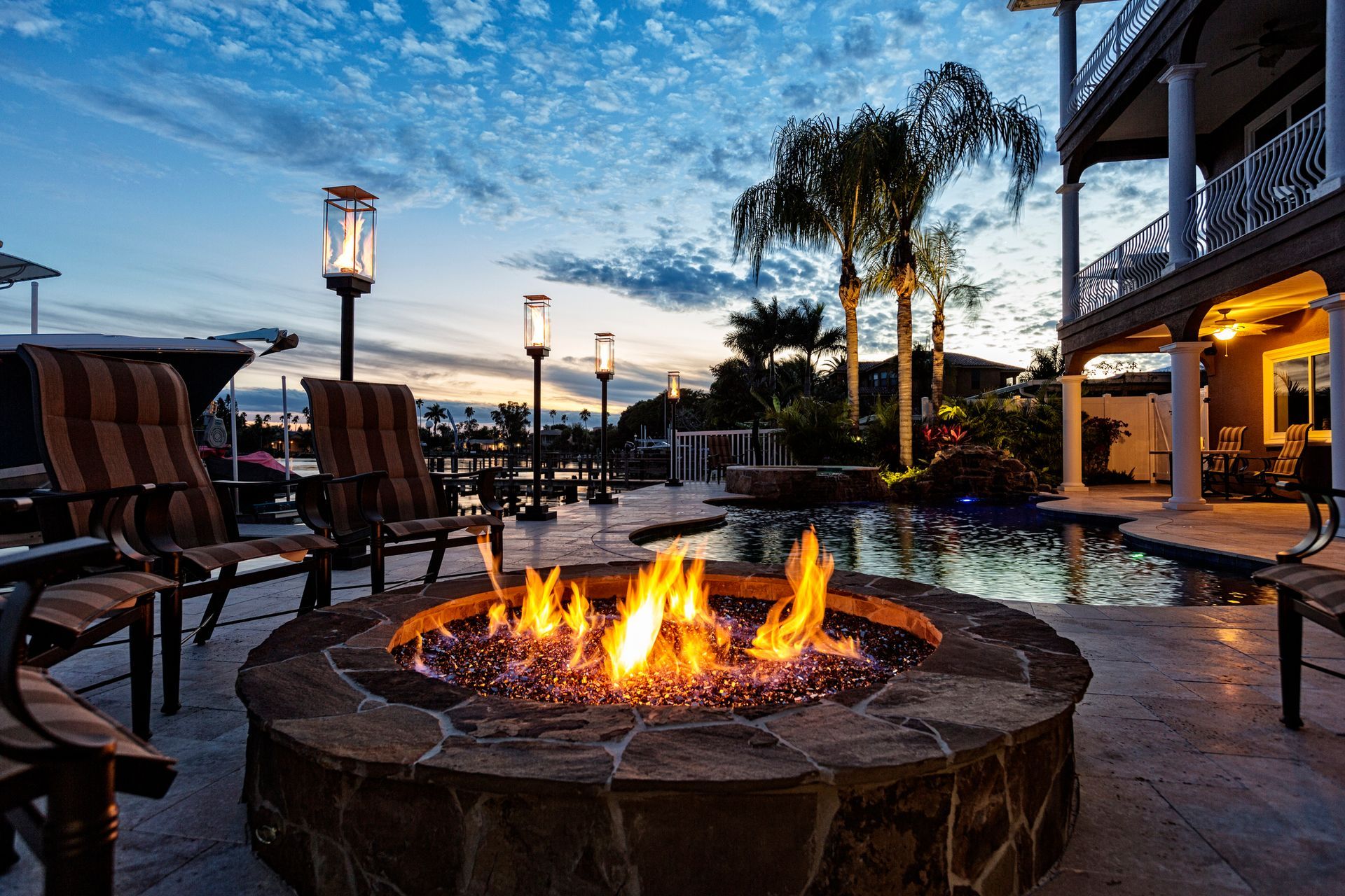 Fire pit with flames, chairs, and pool at dusk, near a building with palm trees.