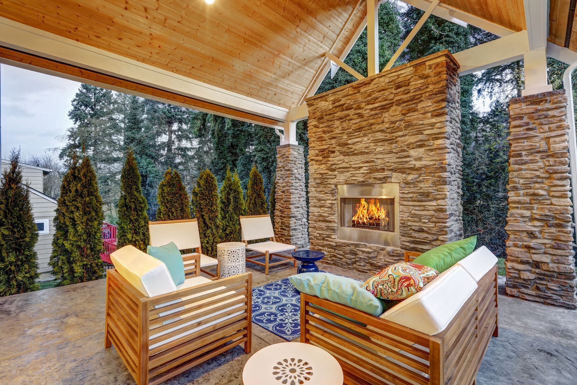 Vaulted patio, stone pillars, teak sofa facing gas fire, white cushions & green pillows.