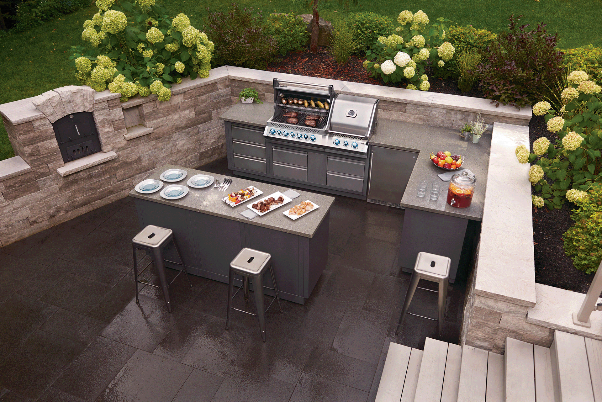 Outdoor kitchen with grill, island, bar stools, and stone wall, surrounded by plants. Outdoor kitchen with grill, island, bar stools, and stone wall, surrounded by plants.