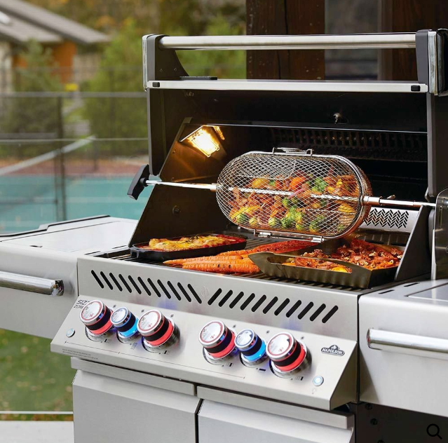 A stainless steel outdoor grill with a rotisserie basket filled with vegetables cooking above other food on the grates.