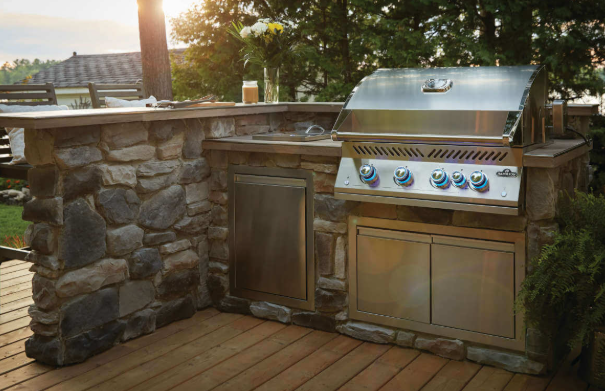 Outdoor kitchen with a stone-clad island, stainless steel grill, and cabinetry on a wooden deck at sunset.