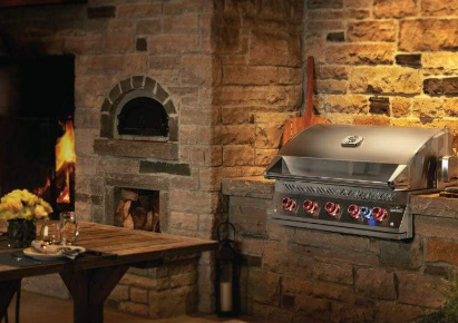 An outdoor kitchen with a stone pizza oven, a lit fireplace, a stainless steel grill, and a rustic wooden dining table.