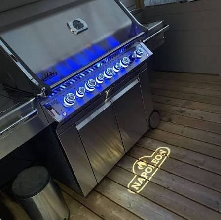 A stainless steel Napoleon gas grill on a wooden deck, with glowing blue control knobs and a projected brand logo on floor.