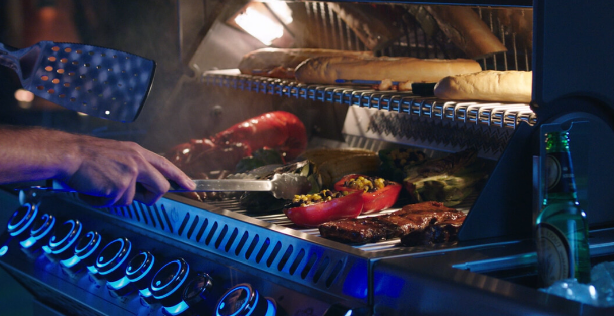A person uses metal tongs to grill vegetables and meat on a large outdoor gas barbecue at night.