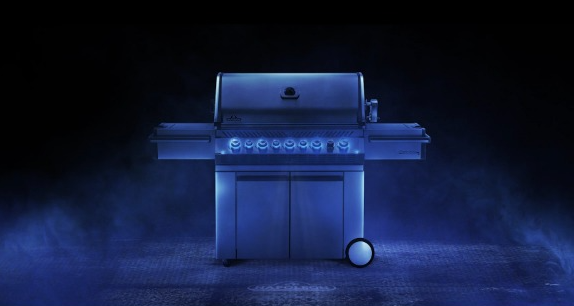 A stainless steel gas grill with illuminated blue knobs stands centered against a dark, moody background with blue fog.