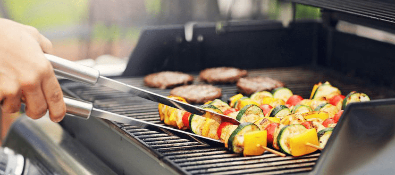 Person using tongs to turn vegetable skewers on a barbecue grill, with burgers in the background.