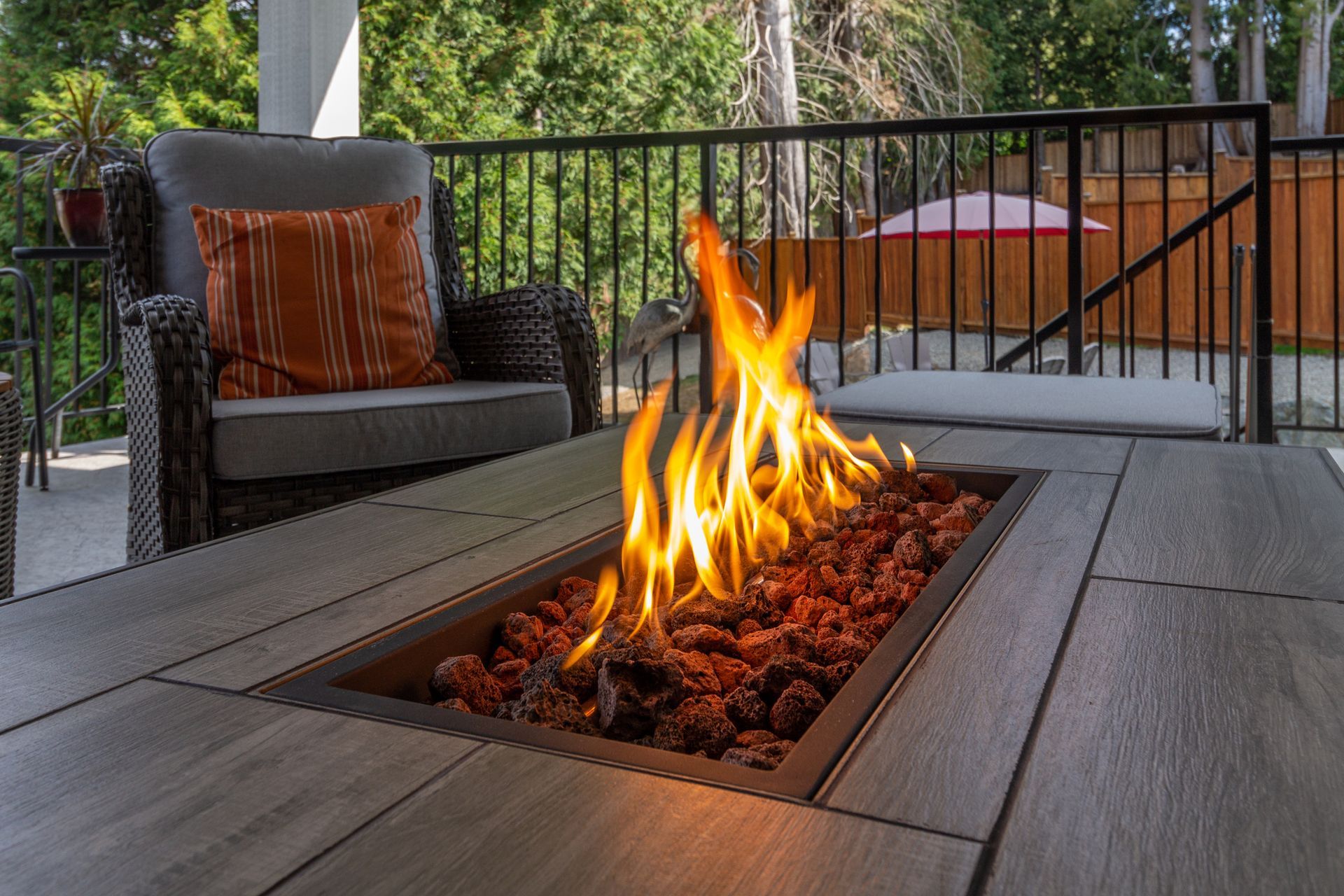 Fire pit with orange flames, surrounded by rocks, on a wooden deck with an armchair and forest background.