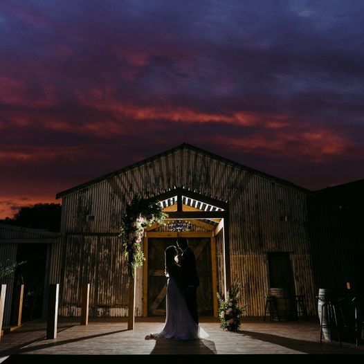 A bride and groom are kissing in front of a barn at sunset.