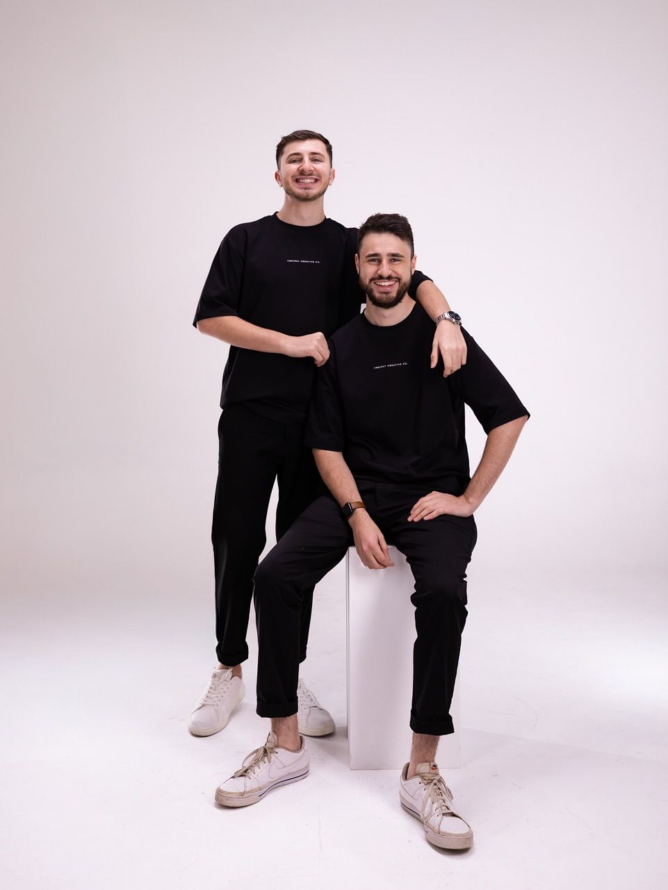 Two men wearing black outfits, posing against a white backdrop. One seated on a white block, other standing.