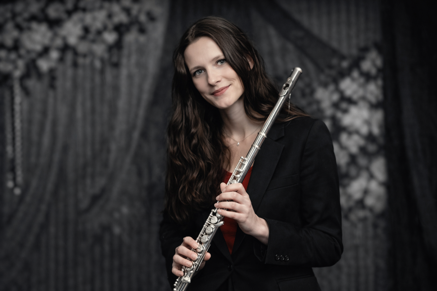 Woman in a black blazer holding a silver flute, smiling, set against a dark, textured backdrop.