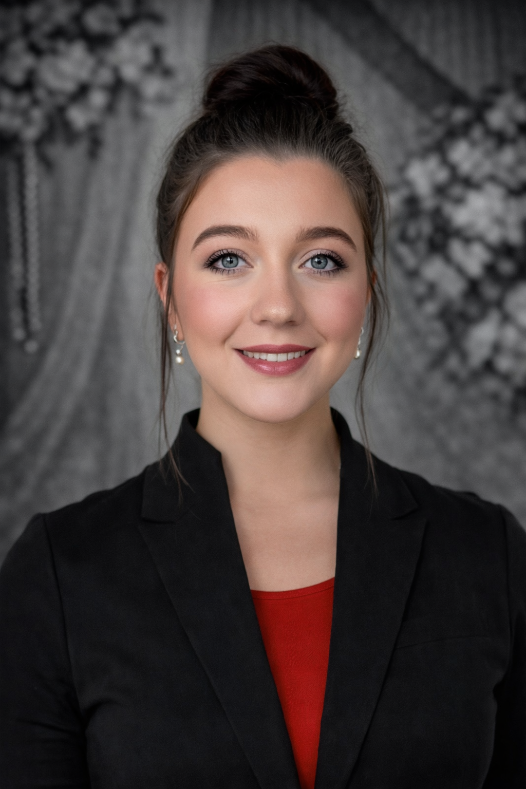 Woman with dark hair in a bun wearing a black blazer and red top smiling, looking at the camera.