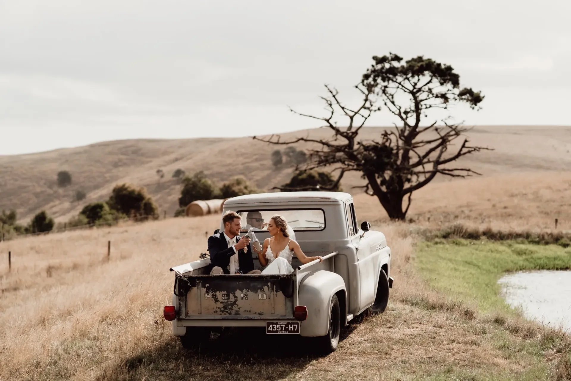 Couple in wedding attire riding in a vintage truck bed, open field with tree in background.