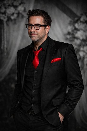 A person in a black suit, vest, and tie with a red necktie and pocket square, posing against a moody backdrop.