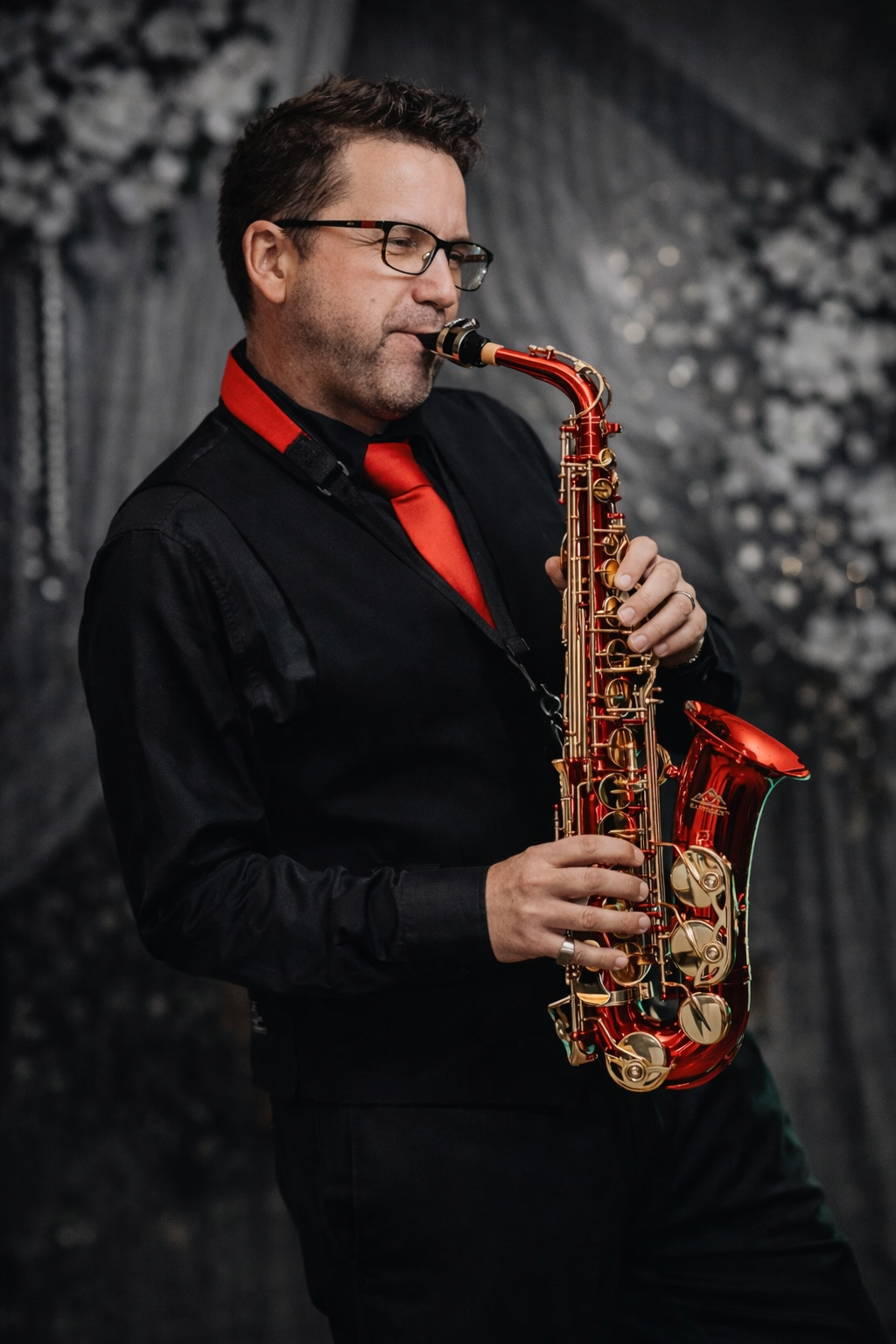 A man in a black shirt and red tie is playing a saxophone.