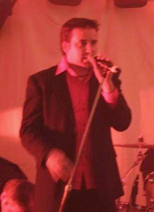 Man in suit jacket and red shirt sings into a microphone. Red stage lighting.