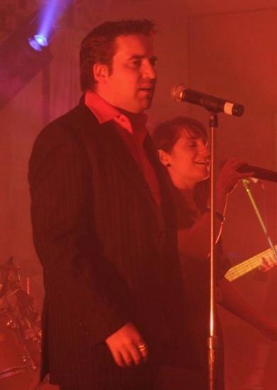 Man in a suit singing into a microphone onstage, red lighting, another person in the background with a microphone.