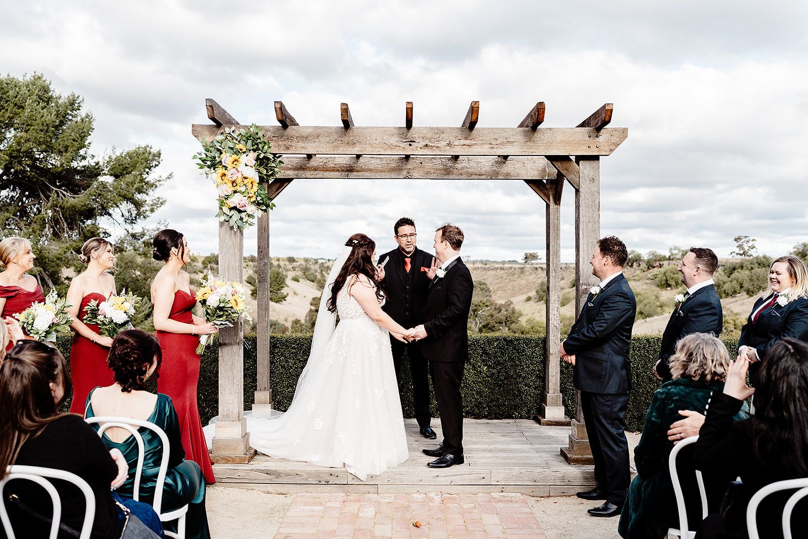 A bride and groom are holding hands during their wedding ceremony under a wooden archway.