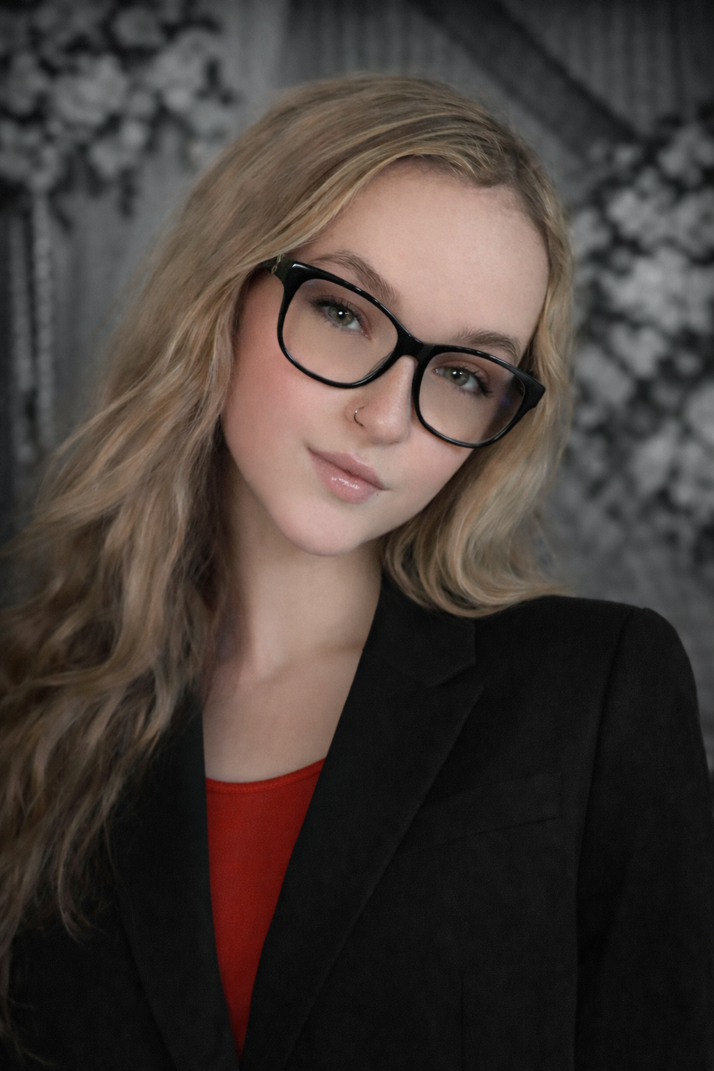 Blond person with glasses in a black blazer and red top, looking at the viewer. Background is a floral print.
