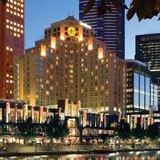 Hotel building at dusk with illuminated facade, near a waterfront in a city, several skyscrapers in the background.