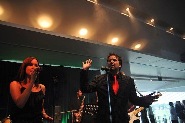 Band performing on stage: woman singing, man in suit with red tie, other musicians visible.