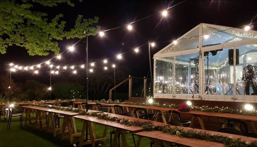 A row of wooden tables are lined up in front of a clear tent at night.