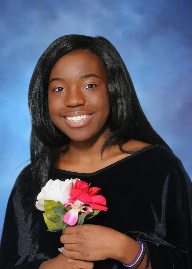 Young Black woman with long, dark hair smiles, holding flowers; against a blue backdrop.