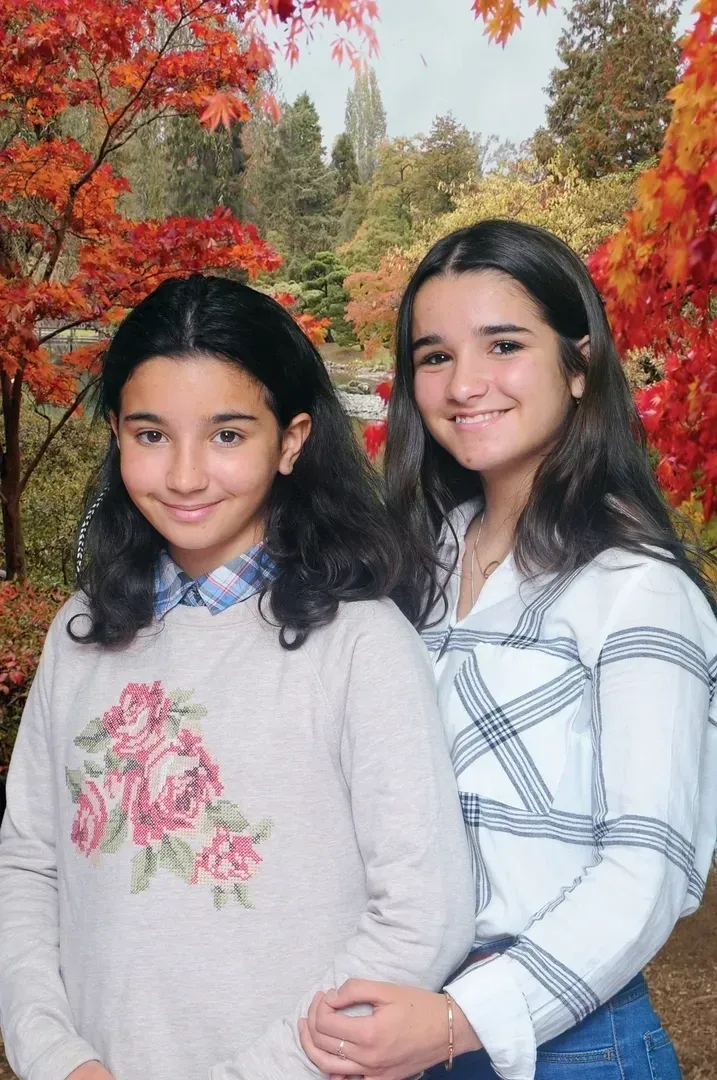 Two young women smiling in front of fall foliage. One wears a floral sweater, the other a plaid shirt.