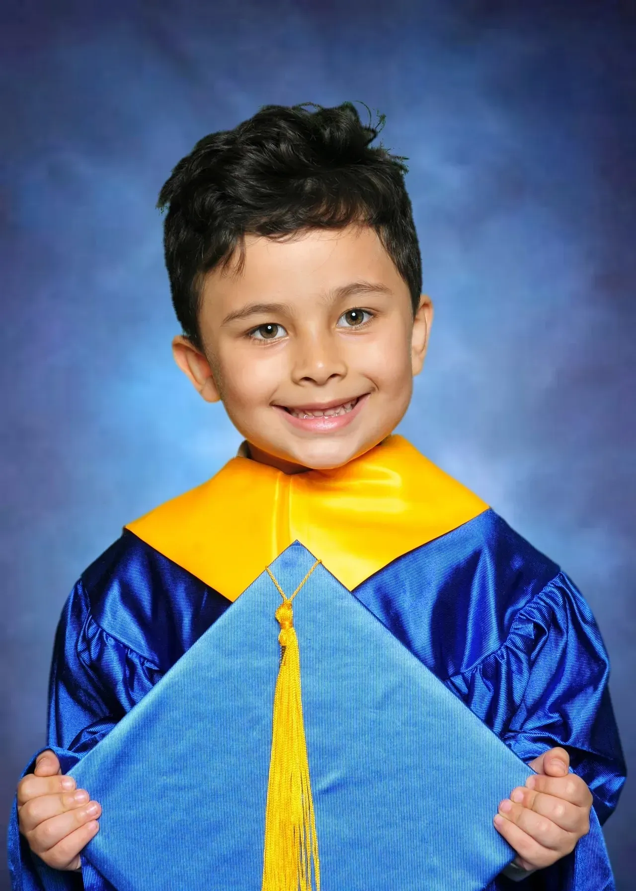 Young boy in blue and gold graduation gown smiles holding his cap.