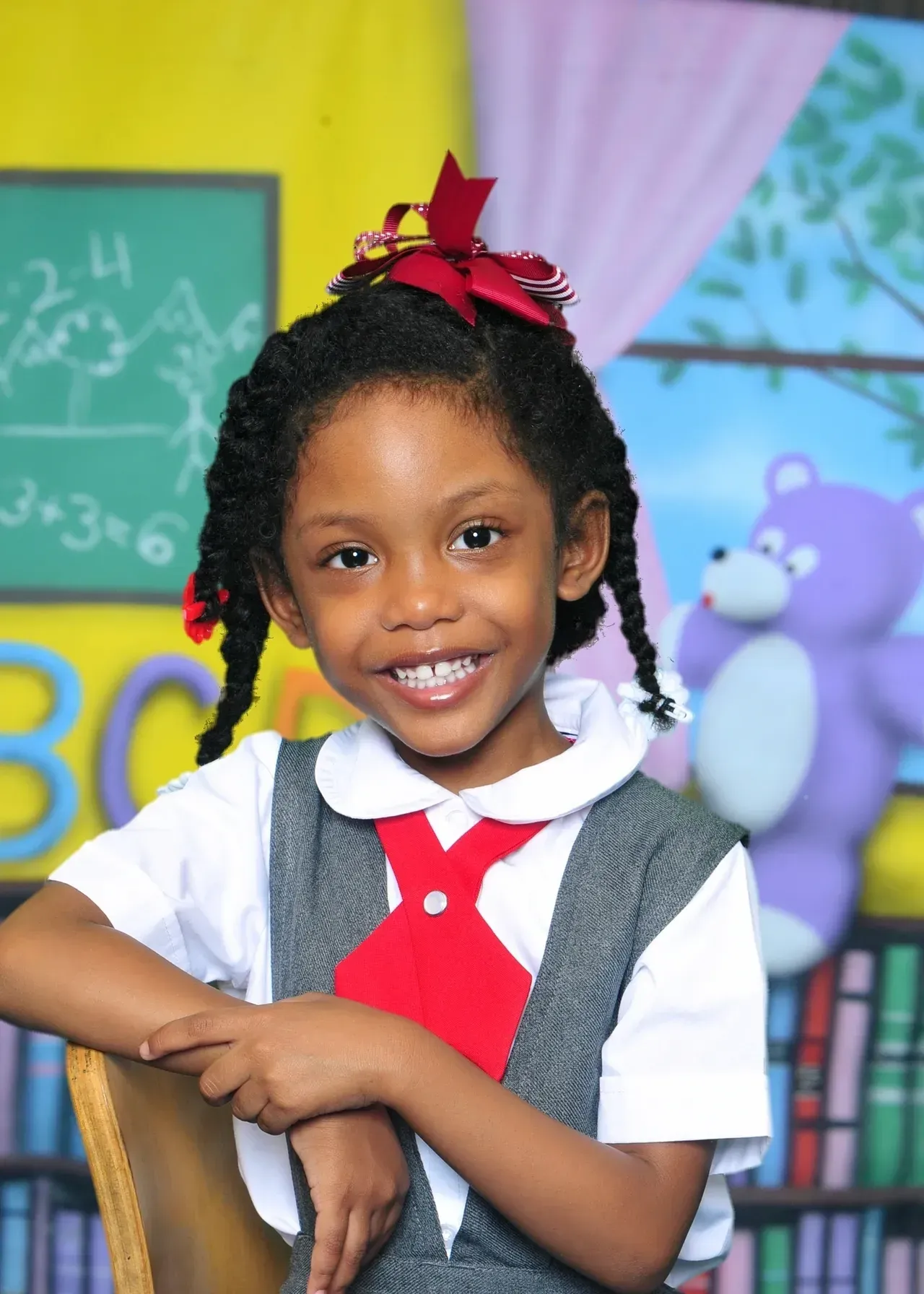 Smiling young girl in school uniform with braided hair, red bow. Classroom backdrop with chalkboard, teddy bear.