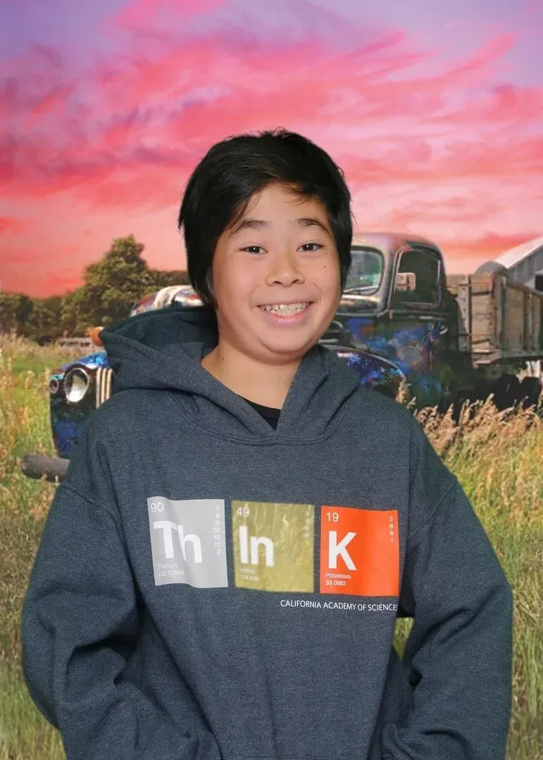 Boy wearing a hoodie with a periodic table design smiles in front of a sunset and old truck.