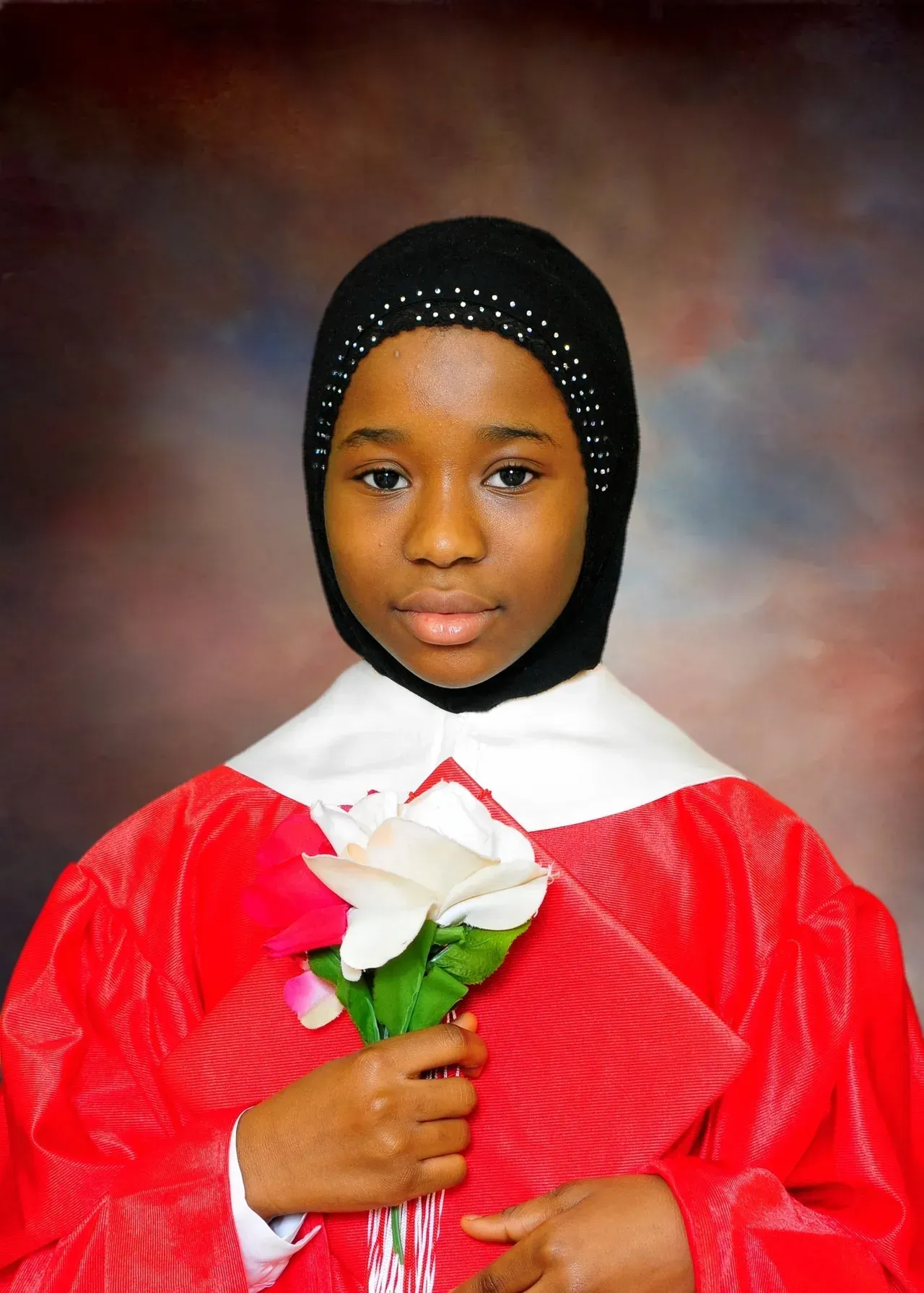 Young Black girl in red graduation gown and black hijab, holding a white flower.