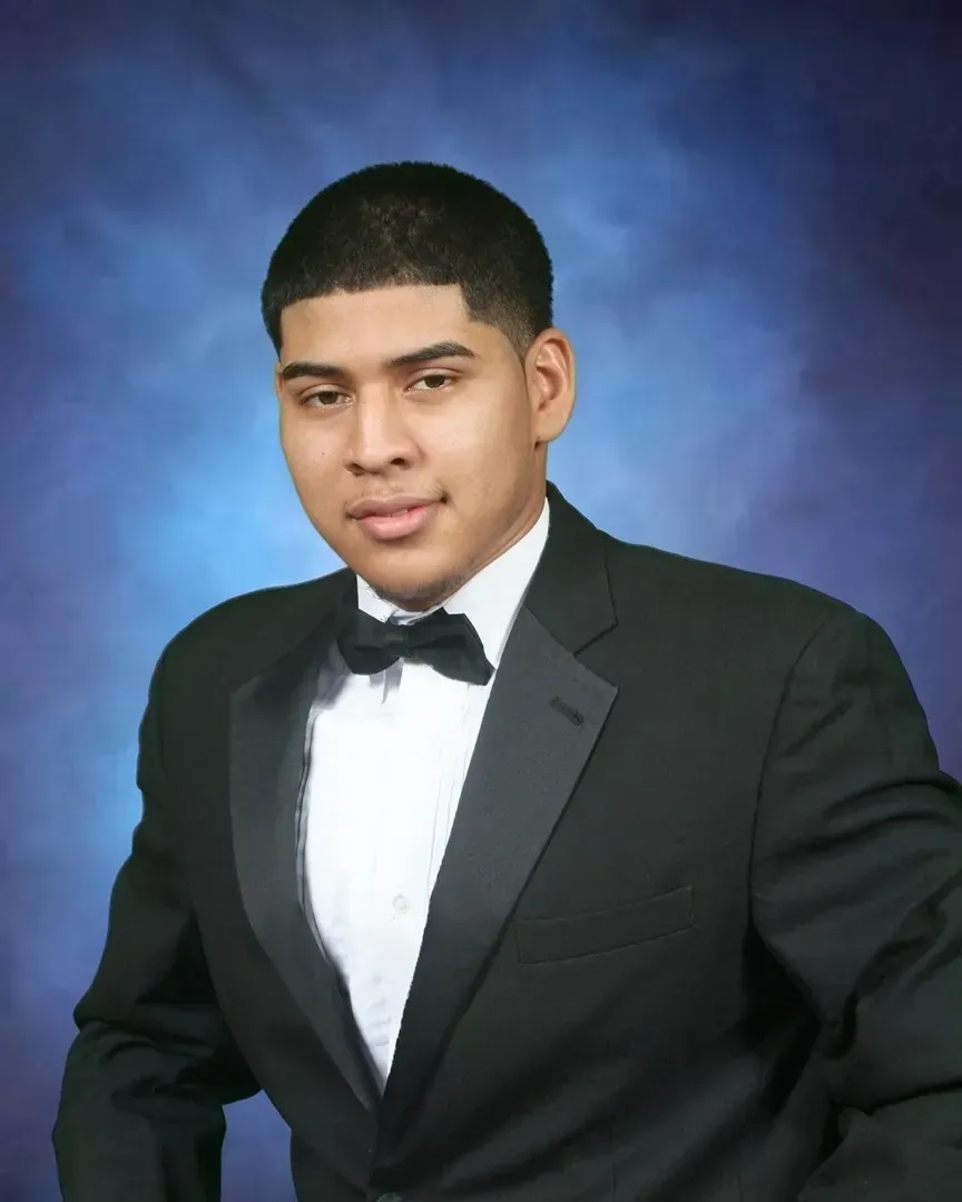 Man in a black tuxedo with a bow tie, against a blue background, smiling.