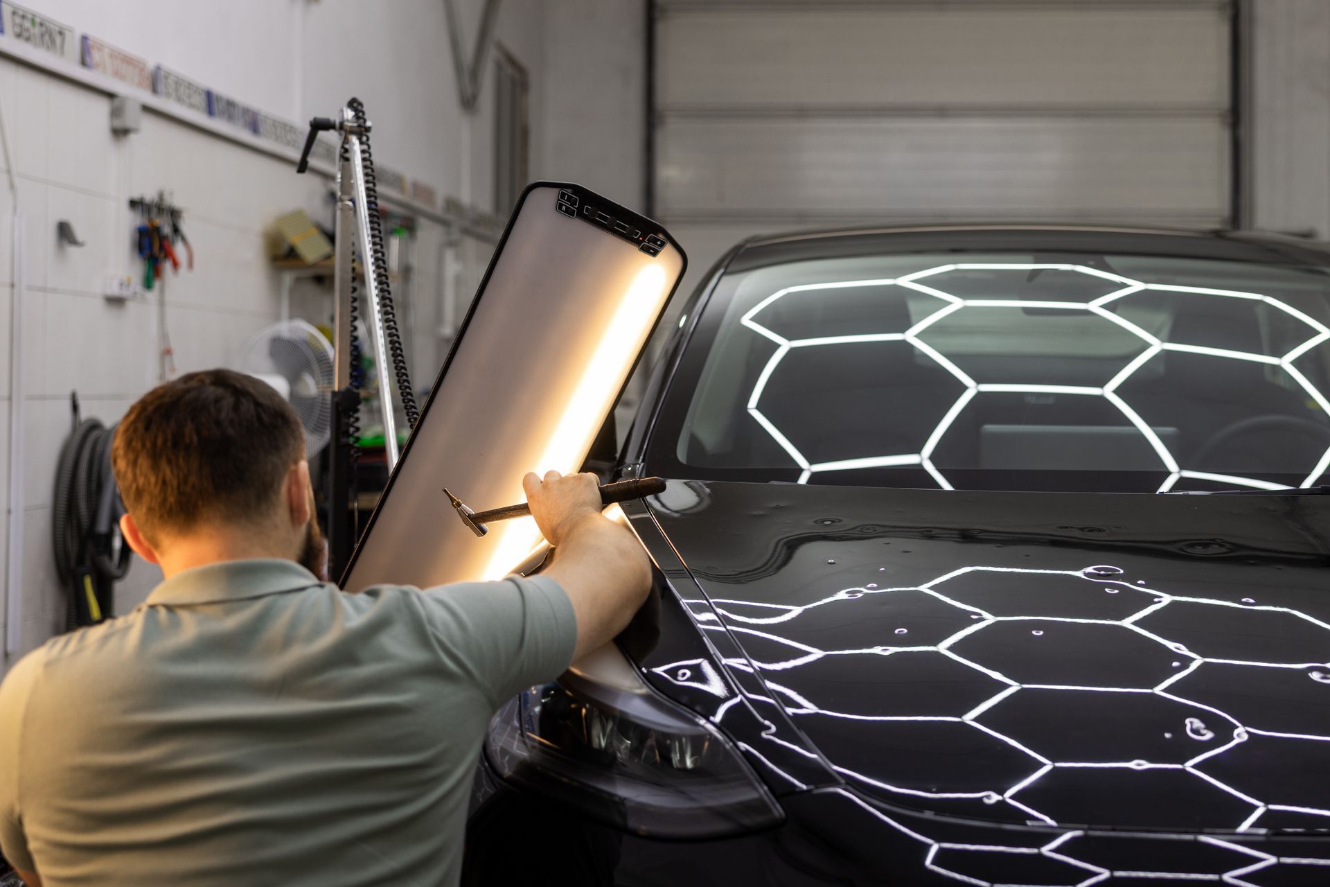 A person using a light to inspect a black car hood for dents in a garage.