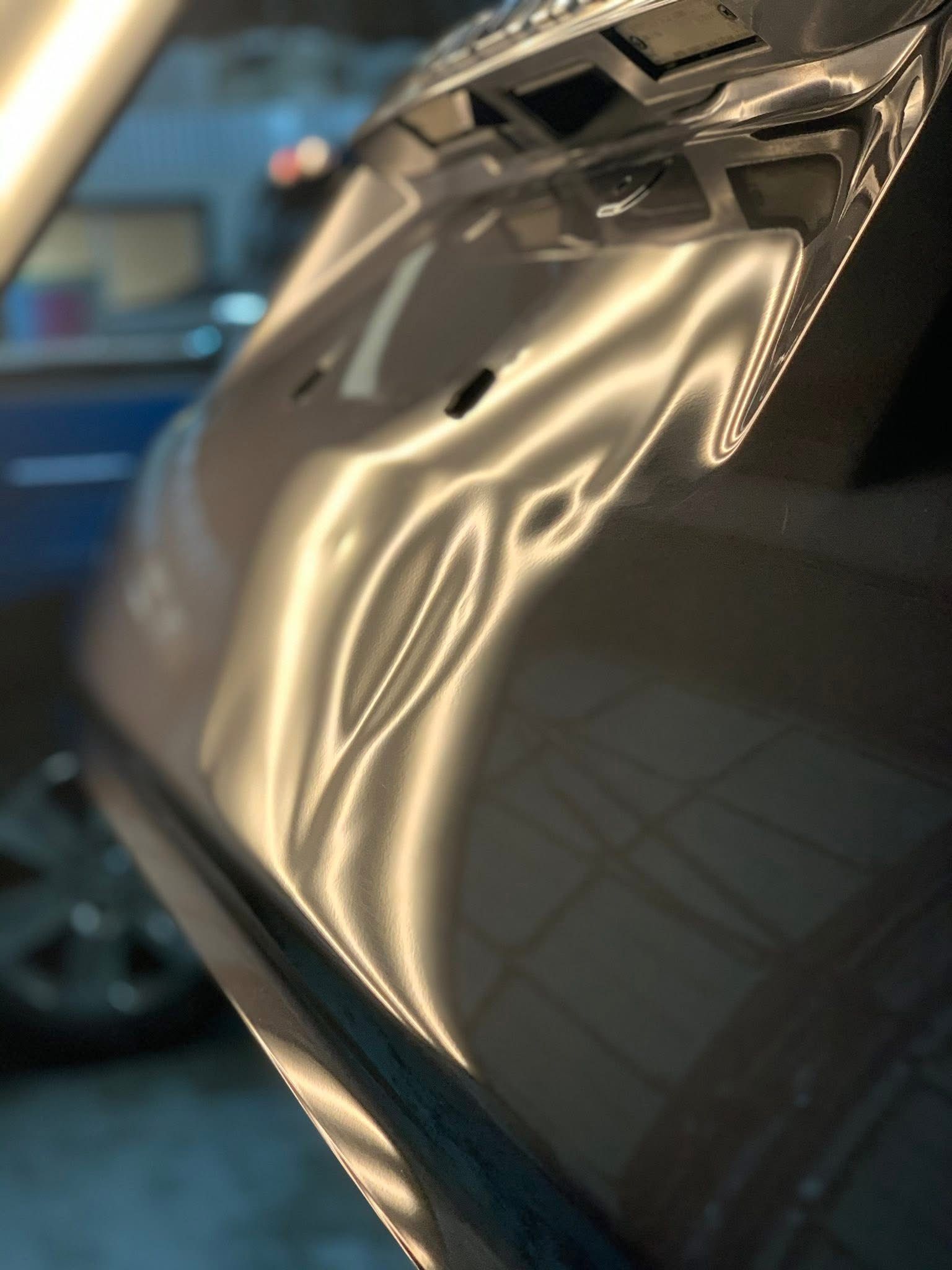 A close-up of a vehicle's dark metallic body panel showing a noticeable dent and surface distortion from light reflection.