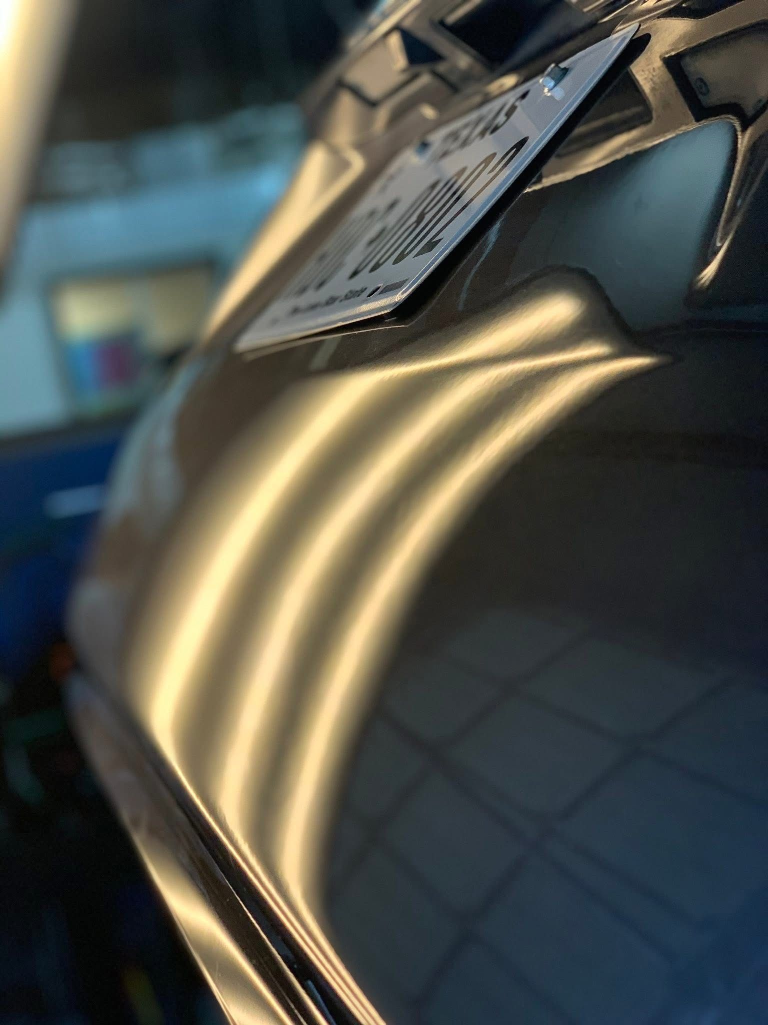 Close-up of a vehicle's rear, featuring a license plate and metallic, curved bodywork reflecting overhead workshop lights.
