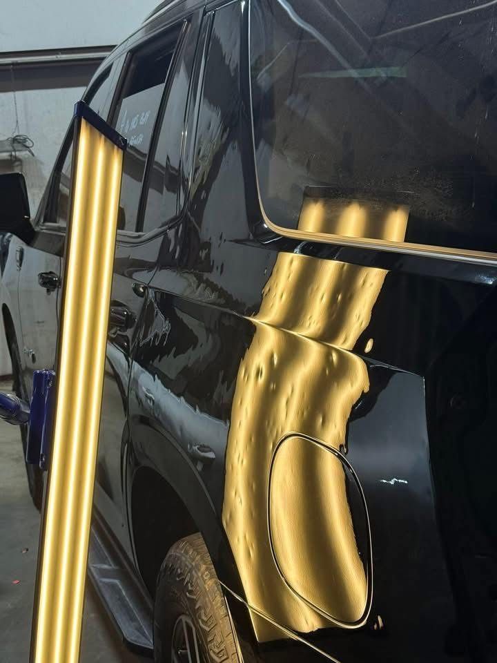 A mechanic uses a bright yellow reflection light board to inspect multiple small dents on a black car's rear panel.
