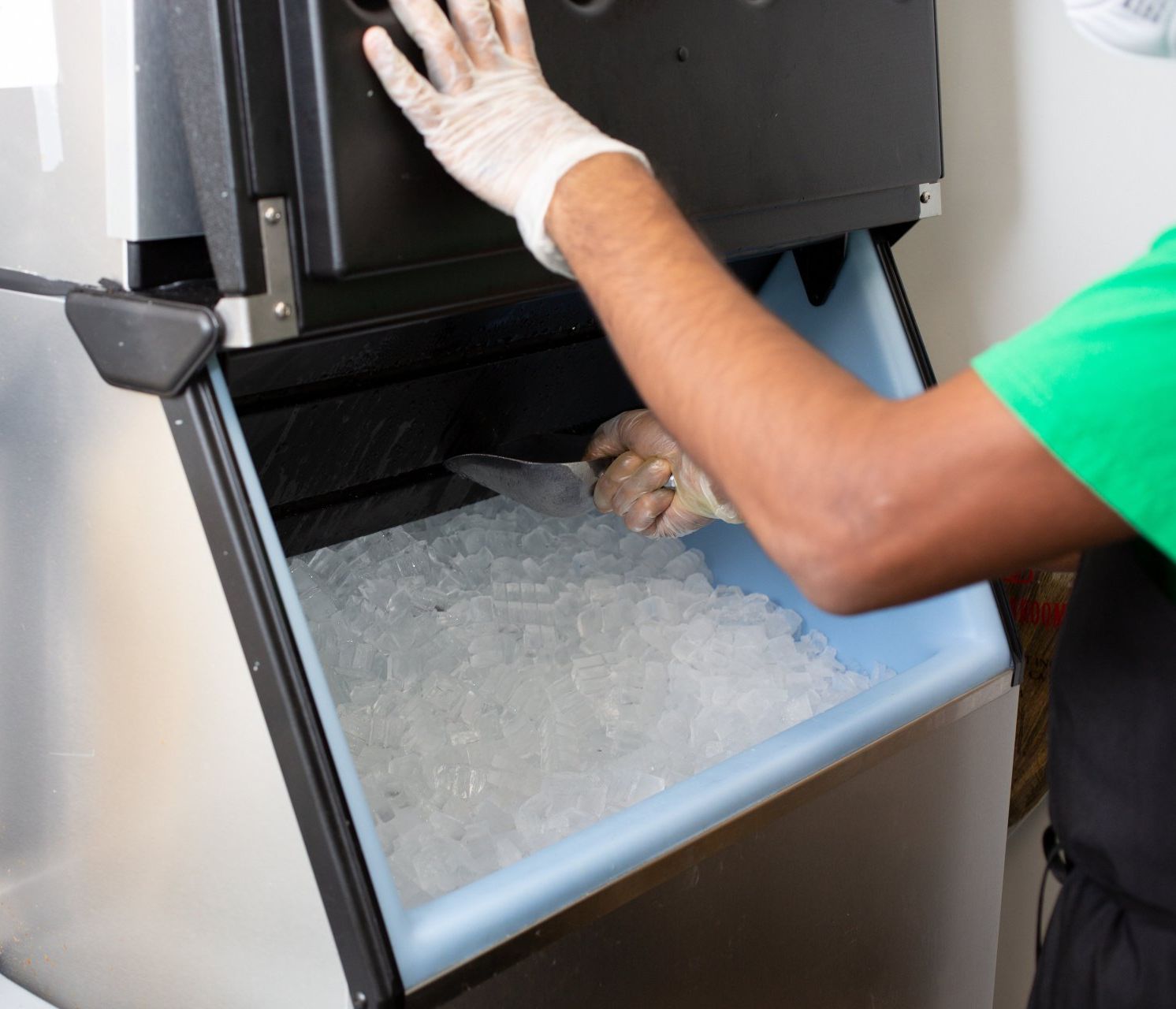 A worker wearing gloves and holding a scoop is seen inside an ice maker, collecting ice cubes into a container.