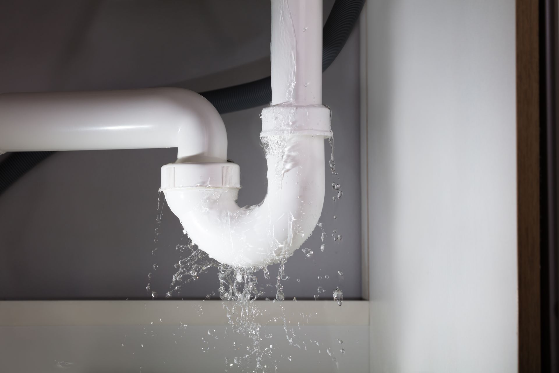 Water leaks from the joints of a white plastic U-shaped drain pipe under a sink.