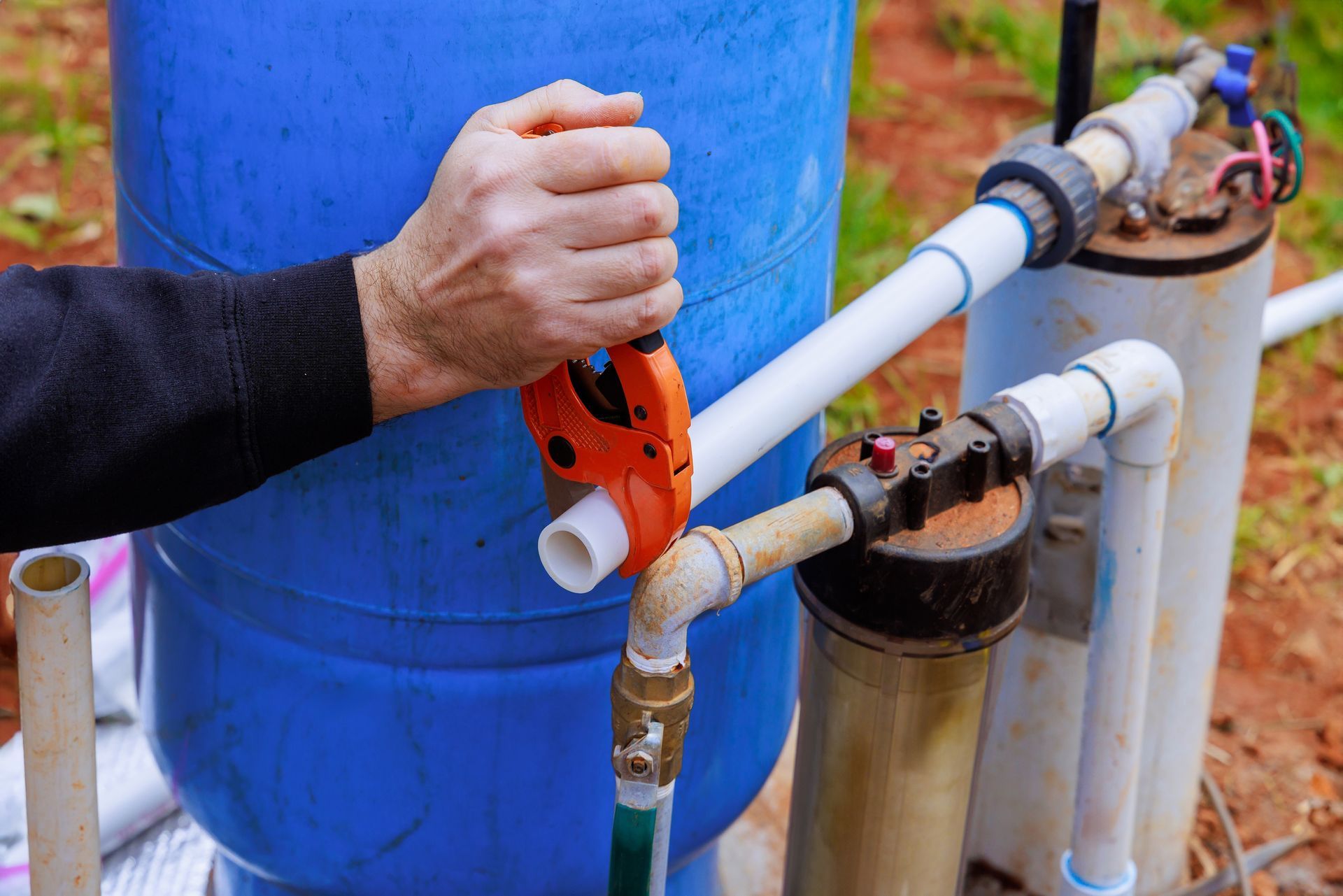 A person uses an orange pipe cutter to cut a white PVC pipe next to a blue water tank and a water filtration system.