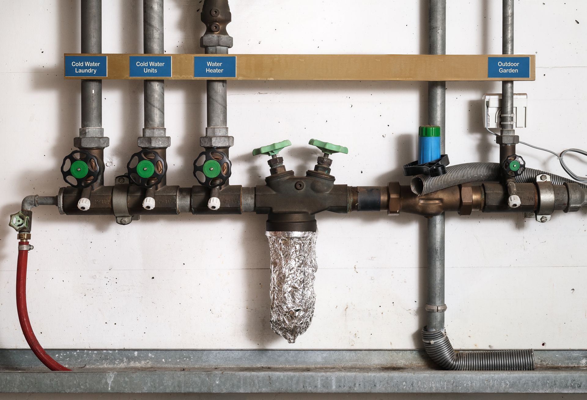 A row of vertical metal pipes mounted on a white wall, featuring green valve handles and a water filter wrapped in foil.