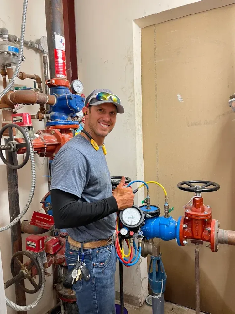 A smiling technician in a gray shirt and cap giving a thumbs up while inspecting a blue valve assembly in a utility room.