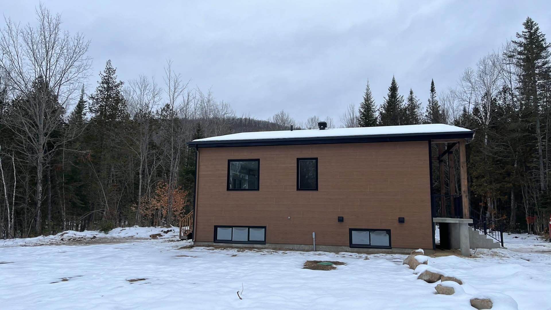 Maison moderne brune dans un cadre boisé et enneigé, sous un ciel nuageux.