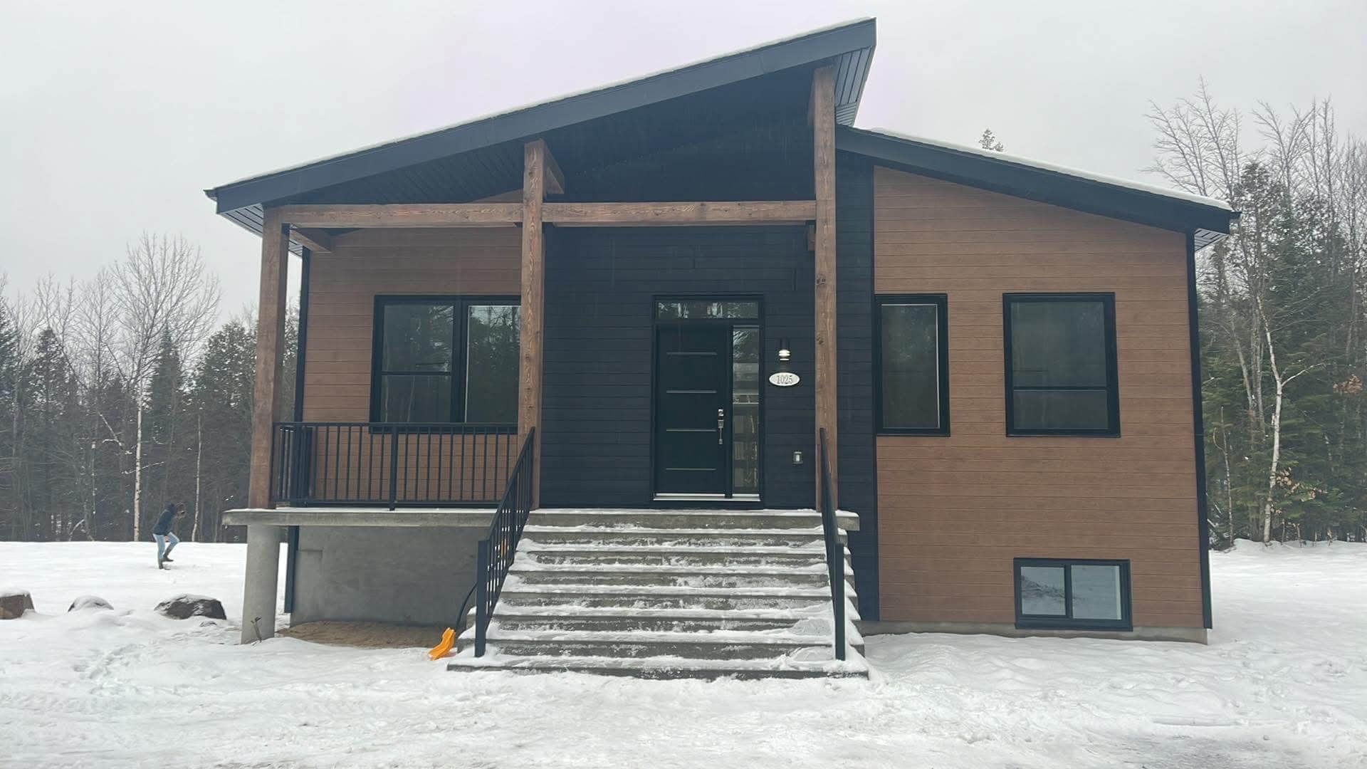 Maison moderne avec bardage noir et marron, marches enneigées menant à la porte d'entrée.