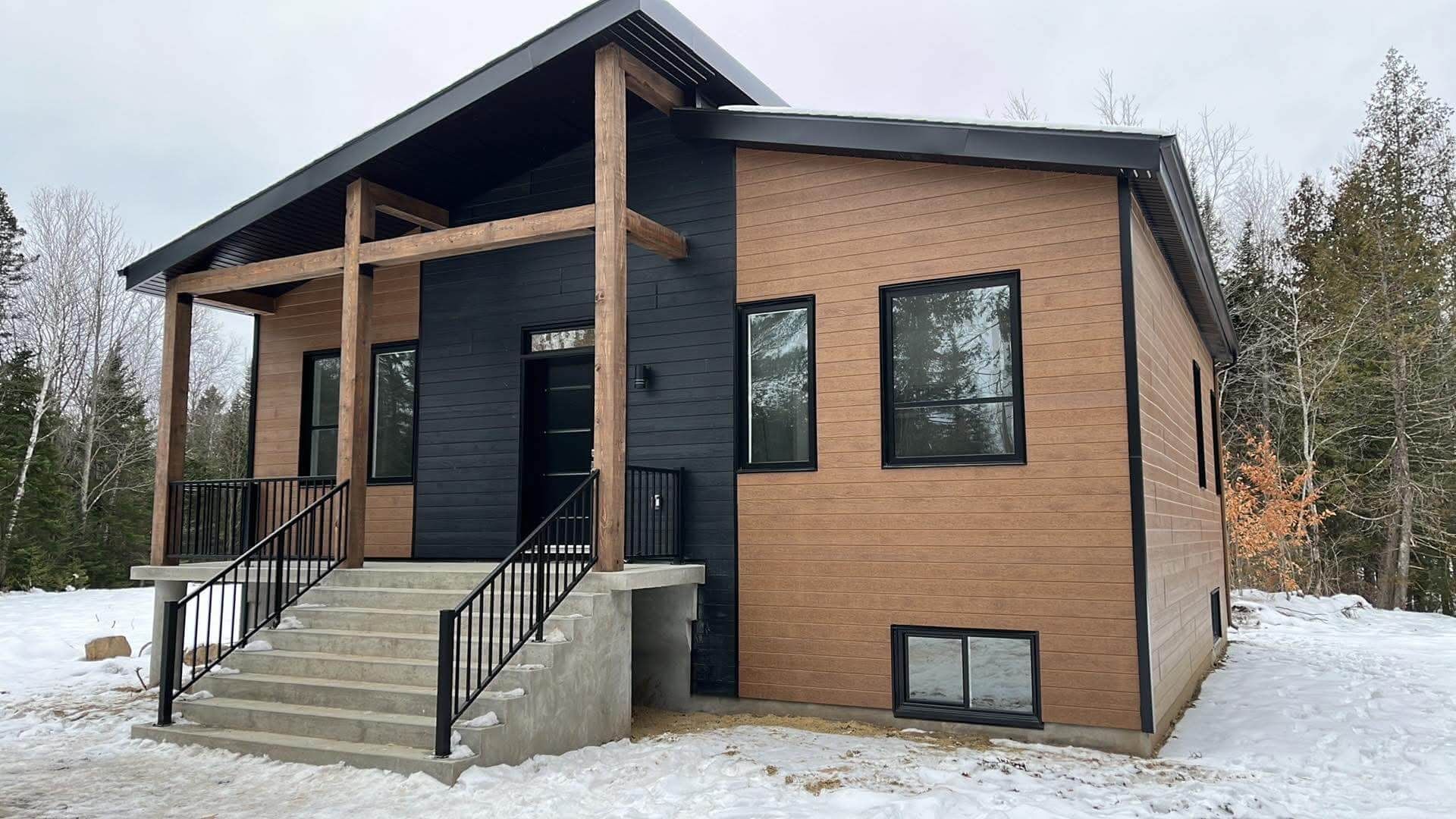Maison moderne avec façade en briques brunes, touches de noir et porche en bois dans un décor enneigé.