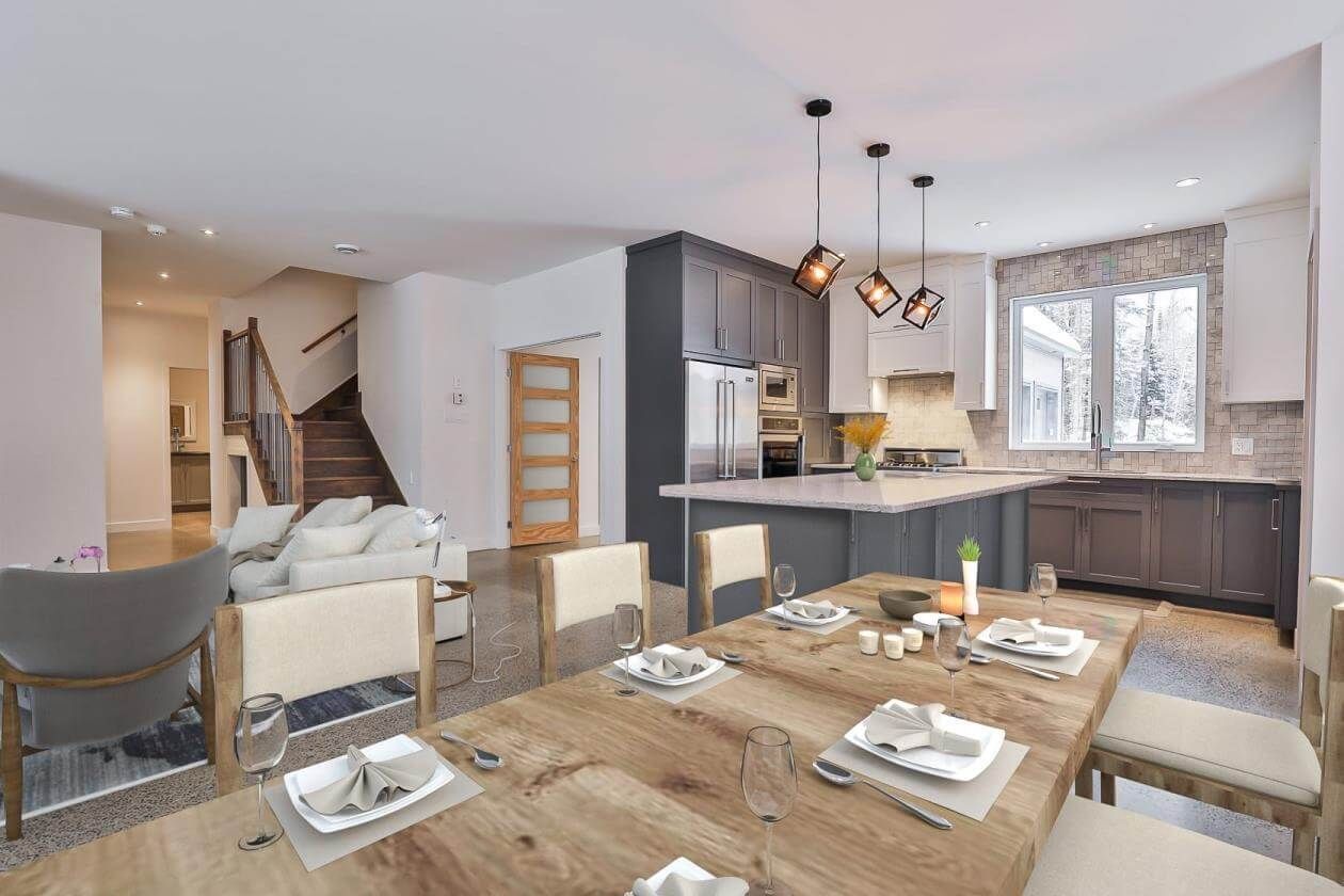 Cuisine et salle à manger ouvertes avec une table en bois dressée pour les repas. Comprend un salon et un escalier.