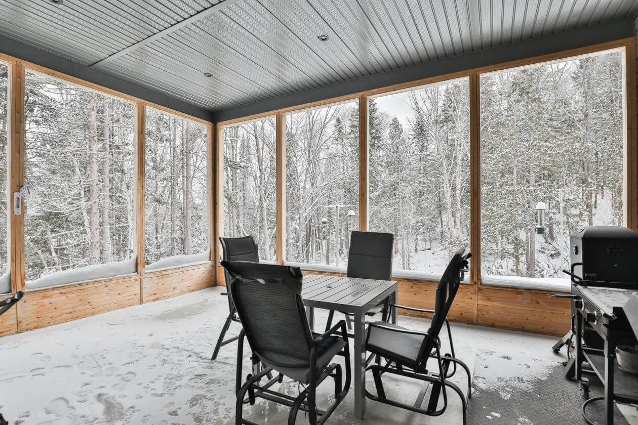 Véranda fermée avec une table et des chaises, donnant sur une forêt enneigée.