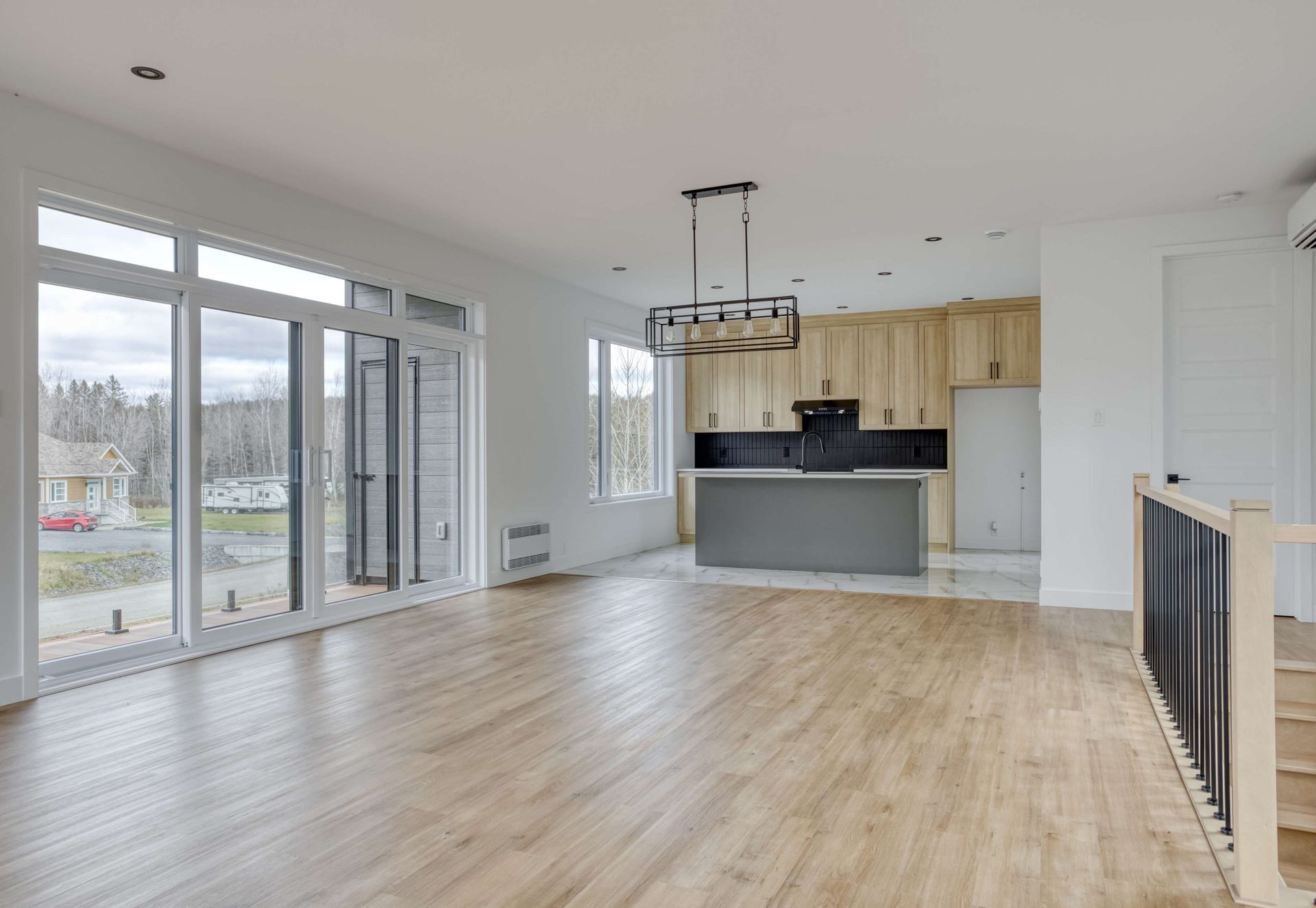 Pièce à vivre spacieuse et vide, avec parquet et cuisine en arrière-plan. Grandes fenêtres, lumière naturelle.