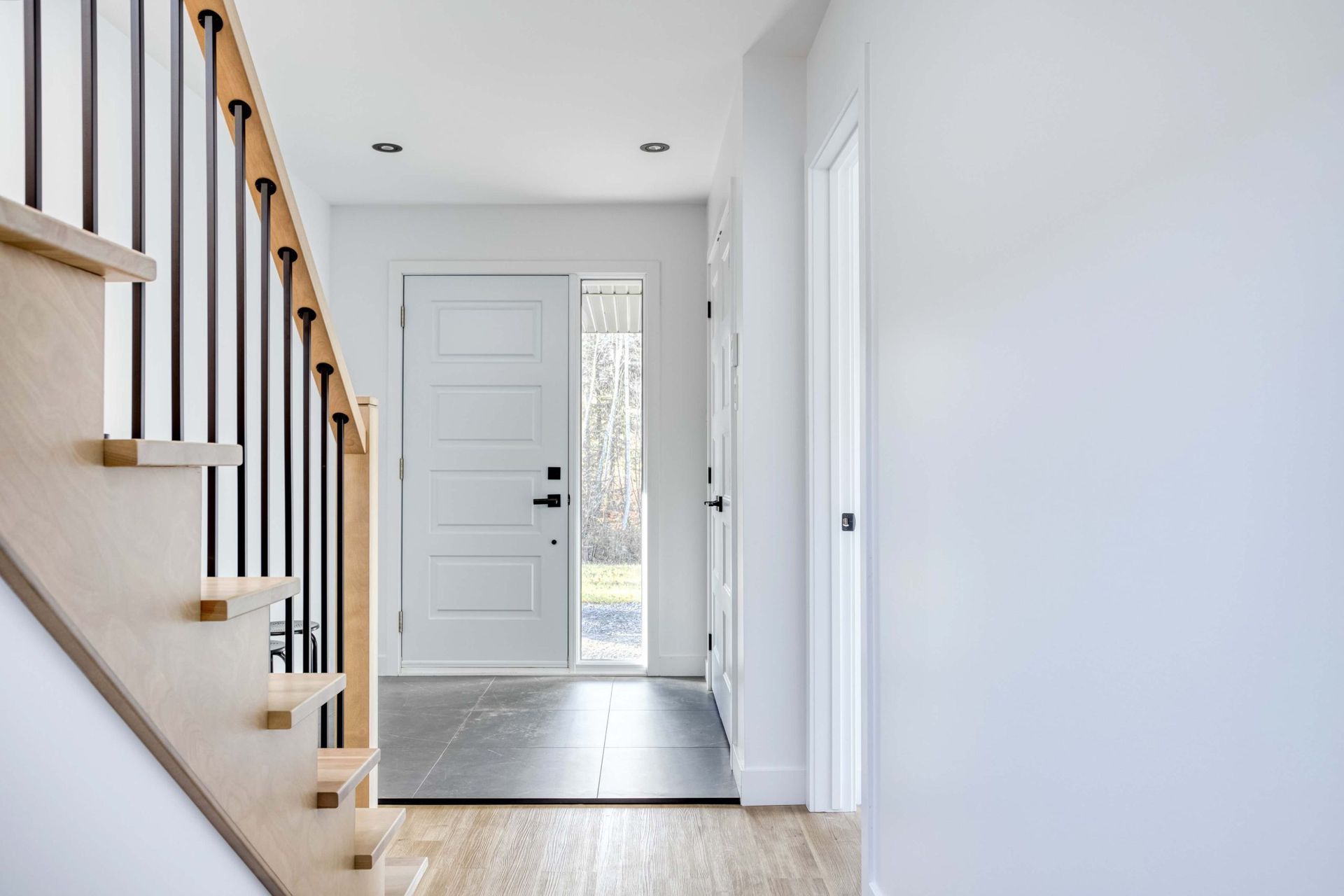 Entrée avec escalier en bois clair, rampe noire, porte blanche et sol gris.